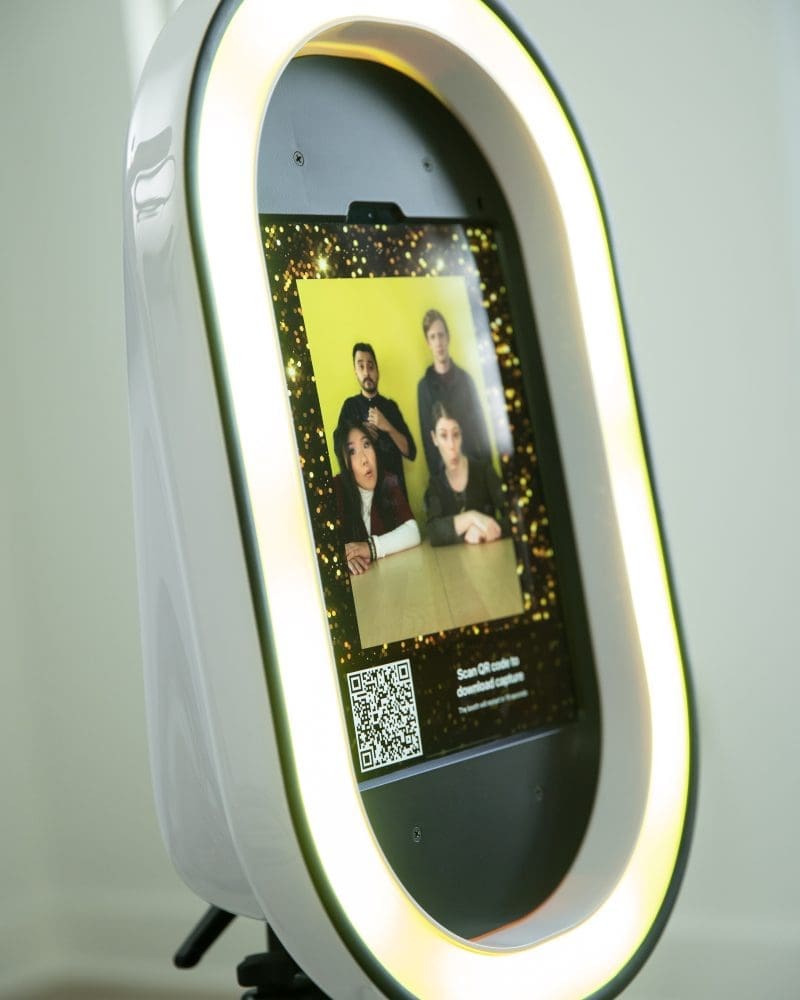 Modern photo booth with LED ring light features a screen displaying a group photo and QR code for easy download.